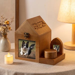 Può includere: Una scatola commemorativa per animali in legno a forma di casa con chiusura in metallo, con una foto di un cane bianco e nero e il testo inciso My darling Deepvoice. Accanto si trova una cornice in legno ad arco più piccola con una foto e una base cilindrica.