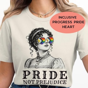 Puede incluir: Una camiseta de color crema con una ilustración de líneas en blanco y negro de una persona con cabello rizado que lleva gafas de sol en forma de corazón con el patrón de la bandera progress pride. El texto PRIDE NOT PREJUDICE aparece en negro.
