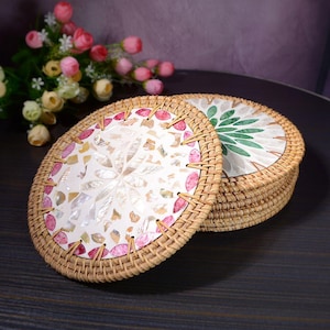 Puede incluir: Un conjunto de posavasos redondos con borde de ratán tejido. Los posavasos presentan un diseño de mosaico blanco con detalles en rosa y beige. Un posavasos tiene un diseño de hojas verdes. Los posavasos están sobre una superficie oscura.