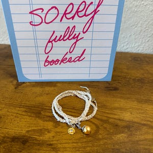 May include: A silver-toned braided cord bracelet featuring a small gold-toned sunflower charm with a central clear stone and a single round gold-toned bead charm. Behind the bracelet is a blue and white card with the text SORRY fully booked.