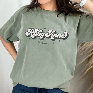 May include: A green t-shirt with a retro-style graphic that says "Ruby Anne" in white and black lettering. The text is surrounded by small stars and the words "Handmade & Co." are printed below.