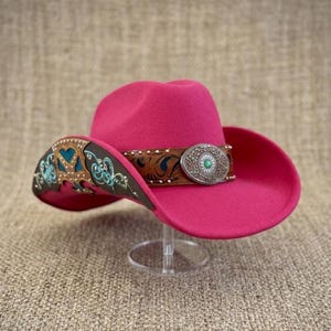 May include: A bright pink felt cowboy hat featuring a brown leather band with intricate floral tooling and turquoise accents. The hat includes a large, oval-shaped silver-toned metal concho buckle at the front with a central green stone inlay.