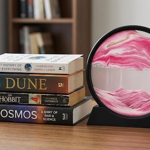May include: A circular sand art frame with a black border and base, containing layers of pink, white, and gold sand. Next to it is a stack of four books titled A Short History of Nearly Everything, Dune, The Hobbit, and Cosmos on a wooden surface.