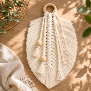 May include: A cream-colored macrame wall hanging shaped like a large leaf, suspended from a smooth wooden ring. The design features intricate knotted cotton cord, two round wooden beads on hanging strands, and a decorative lace ribbon strip.