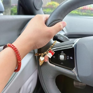 Op de afbeelding: Een rode kralenarmband met een donker verloop aan het uiteinde en een goudkleurige bedelarmband met een wit hart, een rode textuurcilinder en een kleine rechthoekige bedel met rood-witte details en de tekst Drunk Wine.