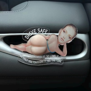 Può includere: Un ornamento decorativo in acrilico trasparente per auto con un'illustrazione stampata di una persona sdraiata in bikini blu. Il testo DRIVE SAFE è stampato sopra, e MY BUTT WOULD BE SO LONELY WITHOUT YOU TOUCHING IT ALL THE TIME sotto.