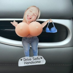 Könnte beinhalten: Ein Auto-Lufterfrischer mit einem Ausschnitt einer Person mit blonden Haaren, die eine blaue Jeans trägt und einen blauen BH hält. Die Figur hat große, runde, pfirsichfarbene Brustaufsätze und eine Basis mit dem Text Drive Safe Handsome.