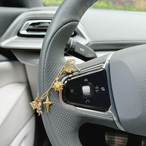 Op de afbeelding: Een goudkleurige bedelarmband met meerdere hangers, waaronder een zon, een vierpuntige ster, een vlinder, een hart en een bloem met een kleine roze steen, hangend over het geperforeerde lederen stuurwiel van een modern voertuig.