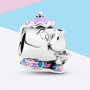 Può includere: Un charm in argento a forma di teiera con un volto, caratterizzato da un coperchio viola e una base decorata con accenti in smalto rosa e blu. La teiera presenta un manico curvo, un beccuccio e un'ampia apertura superiore per gioielli.