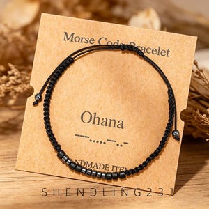 Könnte beinhalten: Ein schwarzes Schnurarmband mit einer Reihe kleiner Metallperlen, die in einem Morsecode-Muster angeordnet sind. Das Armband ist auf einer beigen Karte befestigt, die mit Morse Code Bracelet, Ohana und dem Morsecode-Muster bedruckt ist.