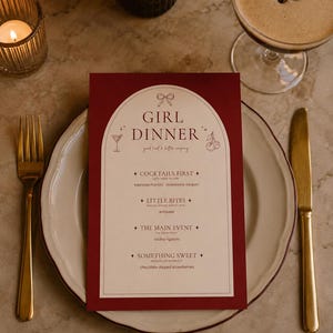 May include: A table setting with a menu for "Girl Dinner." The menu is red with white text and features a bow graphic. A cocktail, a candle, a fork, and a knife are also on the table. The menu lists cocktails, little bites, the main event, and something sweet.