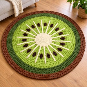 Op de afbeelding: Een rond, gehaakt kleed in de vorm van een doorgesneden kiwi. Het midden is crèmekleurig, omringd door lichtgroen vruchtvlees met stralende witte lijnen en donkerbruine zaadachtige accenten, afgewerkt met een donkerbruine buitenrand.