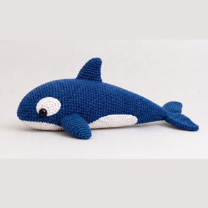 May include: A crochet orca toy featuring a deep blue body with a white underbelly and a white patch around the black eye. The stuffed animal has a prominent dorsal fin, two side pectoral fins, and a wide tail fluke, all crafted in yarn.