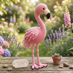 Puede incluir: Una figura amigurumi de flamenco tejida a ganchillo en color rosa con cuello largo, detalles de alas en capas y un pico con punta negra. Junto a ella hay una maceta de terracota con una suculenta y un letrero AMIGURUMI GARDEN HANDCRAFTED.