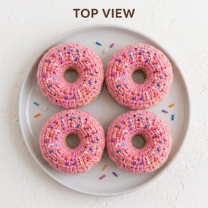 Könnte beinhalten: Vier gehäkelte Donut-Spielzeuge auf einem runden Teller. Jeder Donut hat eine hellbraune Basis mit rosa Glasur und bunten Garnstreuseln in Blau, Orange, Lila und Weiß. Der Text TOP VIEW ist in Braun am oberen Rand des Bildes aufgedruckt.
