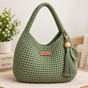 Könnte beinhalten: Eine salbeigrüne gehäkelte Handtasche mit einem einzelnen Henkel und einem rechteckigen braunen Lederetikett, auf dem das Wort Handmade eingeprägt ist. Eine passende grüne Quaste mit einer Holzperle ist seitlich mit einem goldenen Verschluss befestigt.