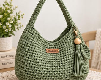 Puede incluir: Un bolso de ganchillo color verde salvia con una sola asa, que presenta una etiqueta rectangular de cuero marrón con la palabra Handmade grabada en el centro. Una borla verde a juego con una cuenta de madera está sujeta al lateral con un cierre dorado.