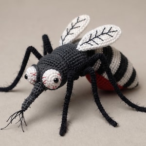Pode incluir: Um brinquedo de mosquito feito em crochê com cabeça e corpo cinza-escuro, asas brancas com veios pretos bordados e pernas longas pretas. O inseto tem olhos brancos grandes com detalhes em fio vermelho, uma probóscide pontiaguda e abdômen listrado.