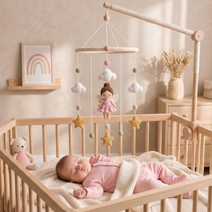 May include: A nursery mobile featuring a central crocheted fairy doll with brown hair and white wings, surrounded by white crocheted clouds, yellow crocheted stars, and small pink and green spheres suspended from a circular wooden frame.