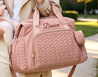 May include: A dusty pink diaper bag featuring a textured herringbone quilted front pocket, gold-tone zippers, and a small matching side pouch. The bag is personalized with the name Danica embroidered in a dark script font on the upper front panel.