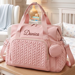 May include: A light pink diaper bag featuring a quilted herringbone pattern on the front pocket and side panels. The bag has two top handles, an adjustable shoulder strap, and a small matching circular pouch. The name Danica is embroidered in brown.