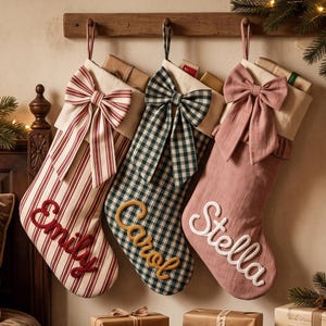 Puede incluir: Tres calcetines navideños colgantes con lazos de tela. El primero es de rayas rojas y blancas con el nombre Emily en rojo. El segundo es de cuadros verdes y blancos con el nombre Carol en amarillo. El tercero es rosa empolvado con el nombre Stella en blanco.