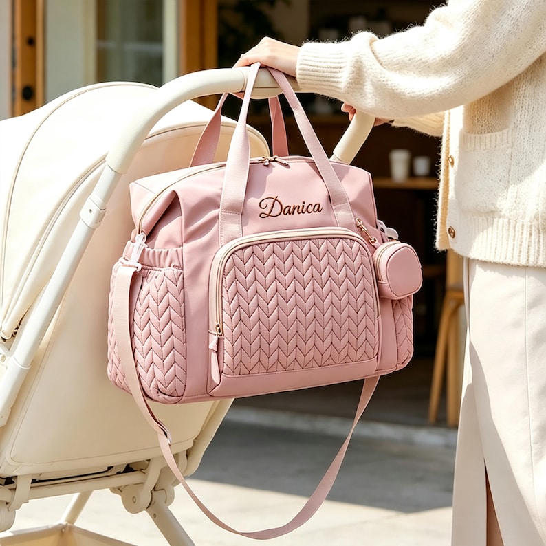 May include: A dusty pink diaper bag featuring a quilted herringbone pattern on the front pocket and side panels. The bag includes two top handles, a detachable shoulder strap, and a small matching pouch. The name Danica is embroidered in dark brown.