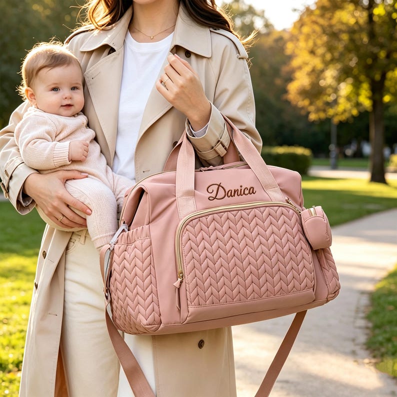 May include: A dusty pink diaper bag featuring a textured herringbone quilted front pocket, gold-tone zippers, and a small matching side pouch. The bag is personalized with the name Danica embroidered in a dark script font on the upper front panel.