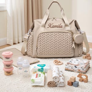 May include: A beige diaper bag with a quilted front panel and the name Kamila embroidered in brown. The bag includes a matching small pouch, two baby bottles, three stackable containers, two packs of wipes, folded cloths, and wooden toys.