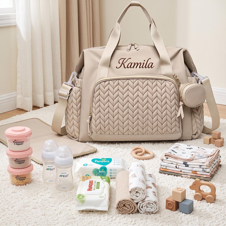 May include: A beige diaper bag with a quilted front panel and the name Kamila embroidered in brown. The bag includes a matching small pouch, two baby bottles, three stackable containers, two packs of wipes, folded cloths, and wooden toys.