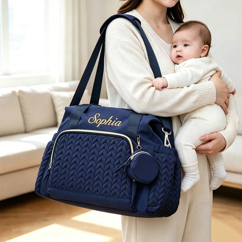 May include: A navy blue diaper bag featuring a quilted chevron pattern on the front pocket, gold-tone zippers, and the name Sophia embroidered in gold script. The bag includes a matching circular pouch attached to the side and sturdy shoulder straps.