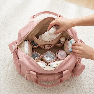 May include: A dusty pink diaper bag with a wide-open top, featuring multiple interior compartments, including insulated pockets, mesh organizers, and a small detachable circular pouch. The bag is filled with baby essentials like diapers, a bottle, and a cloth.