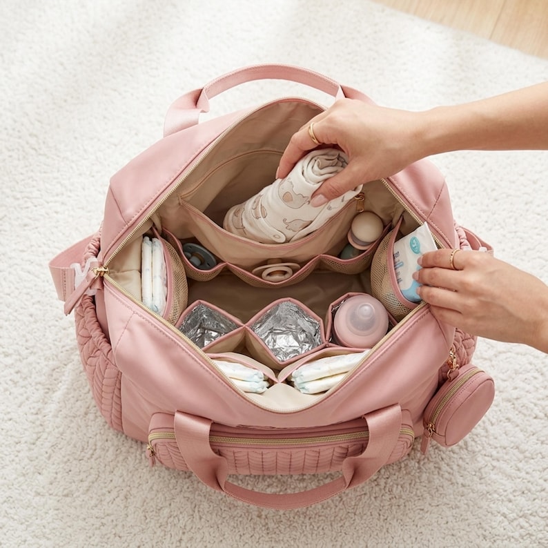 May include: A dusty pink diaper bag with a wide-open top, featuring multiple interior compartments, including insulated pockets, mesh organizers, and a small detachable circular pouch. The bag is filled with baby essentials like diapers, a bottle, and a cloth.