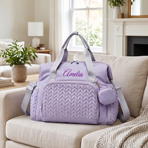 May include: A lavender diaper bag featuring a quilted herringbone pattern on the front pocket and side panels. The bag has light gray straps, silver-tone zippers, and a small detachable circular pouch. The name Amelia is embroidered in purple script.
