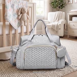May include: A light gray diaper bag with a quilted front panel, gold-tone zippers, and the name Rosalie embroidered in white. The bag features a small detachable pouch and measures 14.2 inches wide, 11.8 inches high, and 7.1 inches deep.