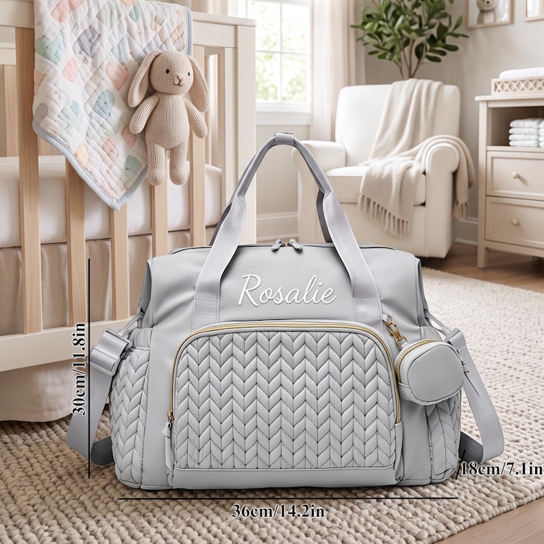 May include: A light gray diaper bag with a quilted front panel, gold-tone zippers, and the name Rosalie embroidered in white. The bag features a small detachable pouch and measures 14.2 inches wide, 11.8 inches high, and 7.1 inches deep.