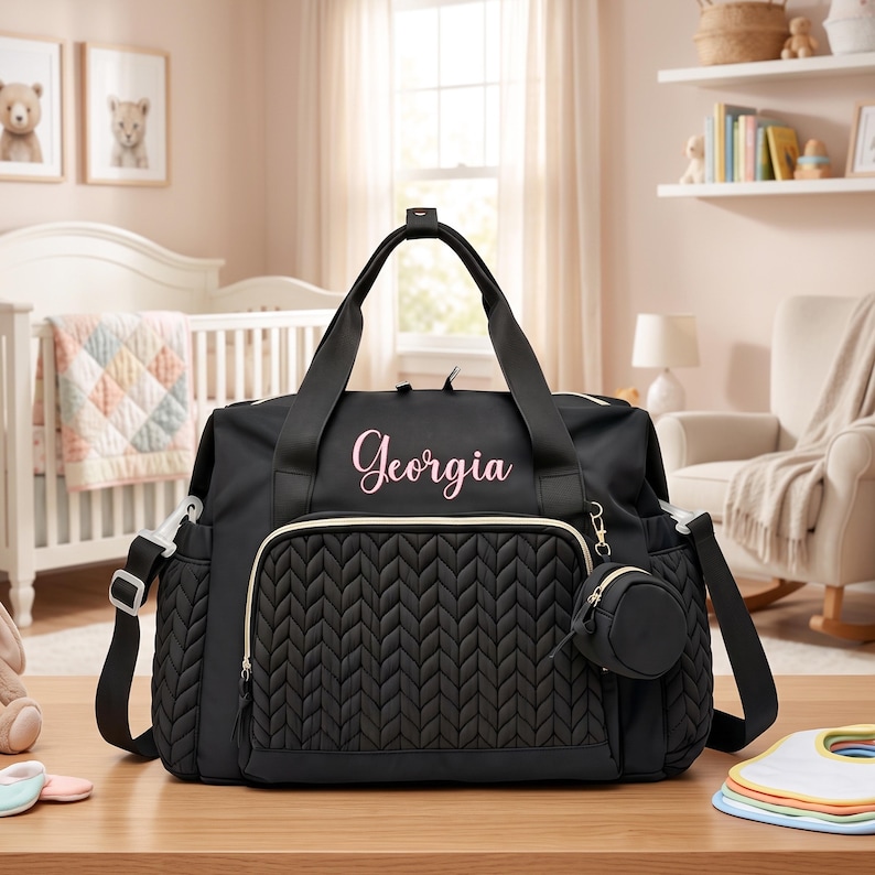 May include: A black diaper bag featuring a quilted chevron pattern on the front pocket, gold-tone zippers, and the name Georgia embroidered in pink script. The bag includes a detachable round black pouch and adjustable black shoulder straps.
