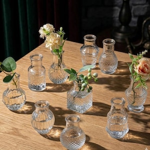 May include: A collection of nine small, clear glass vases with various embossed textures and shapes. Some vases contain artificial flowers and greenery, while others are empty. The vases feature patterns like diamond, ribbed, and crosshatch designs.