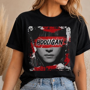 Puede incluir: Una camiseta negra de cuello redondo con un estampado gráfico cuadrado. El diseño muestra un retrato en blanco y negro con un trazo de pincel rojo sobre los ojos que contiene el texto negro HOOLIGAN, rodeado de pintura roja y blanca desgastada.