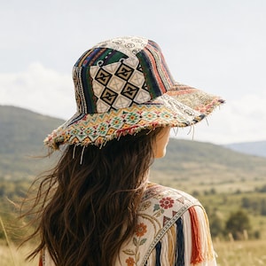 May include: A bucket hat featuring a patchwork design of geometric patterns in black, white, red, green, and blue with a frayed brim. The hat is worn with a cream-colored garment adorned with floral embroidery and long, multi-colored hanging tassels.