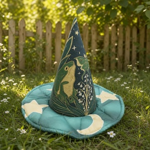 May include: A conical hat in dark teal fabric featuring a printed illustration of a green frog, a moth, and white lily of the valley flowers. The wide, quilted brim is decorated with pale yellow stars and a crescent moon pattern.