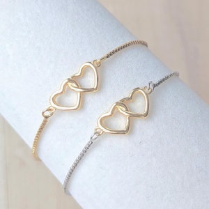 May include: Two delicate gold heart charm bracelets on a white display stand. The charms are intertwined and feature a simple, elegant design.