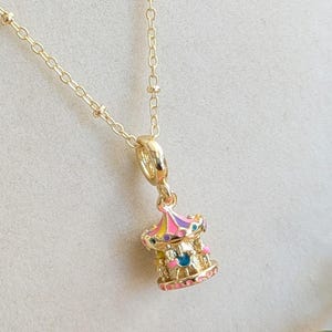 May include: A gold necklace with a carousel pendant. The pendant has a colourful design with pink, yellow, and blue accents. The necklace is displayed on a white mannequin and has a delicate chain.