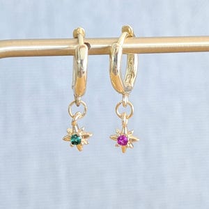 May include: Gold-coloured hoop earrings with starburst charms. One charm has a green gemstone, the other a pink gemstone. The earrings are displayed on a gold-coloured earring stand against a light blue background.