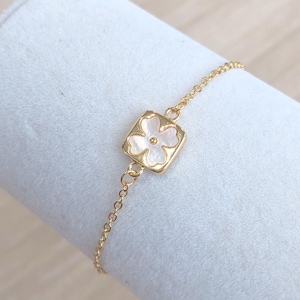 May include: A delicate gold chain bracelet with a square-shaped pendant featuring a white flower design.