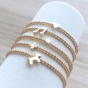 May include: A set of five gold beaded bracelets with different charms, including a heart, a music note, a crown, a cross, and a dog. The bracelets are stacked on top of each other on a white surface.