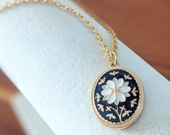 May include: Gold-tone necklace with an oval pendant featuring a white flower design on a black background. The pendant is framed with a gold border and small gold accents. The chain is delicate and gold.