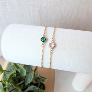 May include: Two delicate gold chain bracelets, each featuring a single, round gemstone. One bracelet has a teal-colored stone, the other a pink stone. The bracelets are displayed on a white, cylindrical surface.