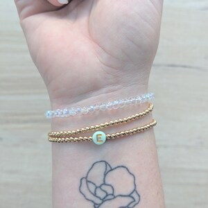 K&ouml;nnte beinhalten: Ein Stapel aus drei Armb&auml;ndern an einem Handgelenk. Das obere Armband besteht aus klaren, schimmernden Perlen. Das mittlere Armband besteht aus goldenen Perlen. Das untere Armband besteht aus goldenen Perlen mit einer hellgr&uuml;nen Perle, auf der ein goldener Buchstabe "E" steht.