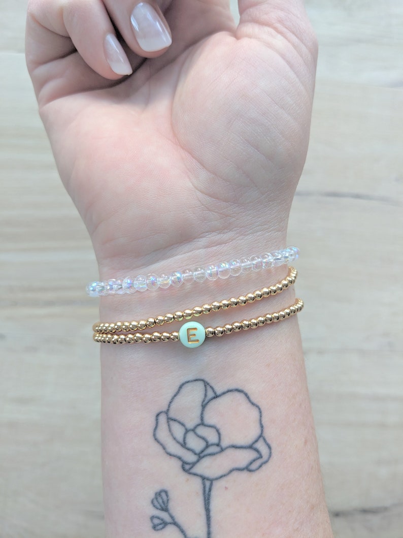 K&ouml;nnte beinhalten: Ein Stapel aus drei Armb&auml;ndern an einem Handgelenk. Das obere Armband besteht aus klaren, schimmernden Perlen. Das mittlere Armband besteht aus goldenen Perlen. Das untere Armband besteht aus goldenen Perlen mit einer hellgr&uuml;nen Perle, auf der ein goldener Buchstabe "E" steht.